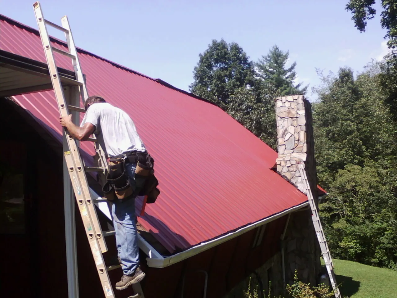 Roofer on a ladder beside a red metal roof and stone chimney — Soffit & Fascia Repair by Roofing Brothers in Lake Winola